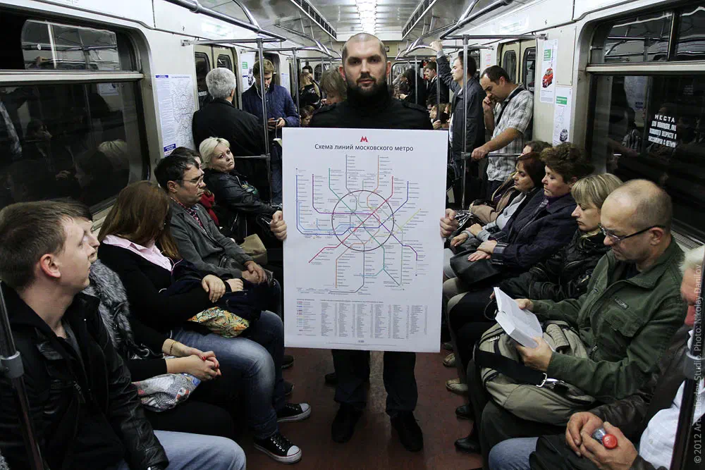 На рисунке изображен человек, стоящий посреди поезда метро, держащий карту перед толпой людей сидящих и стоящих на сиденьях.