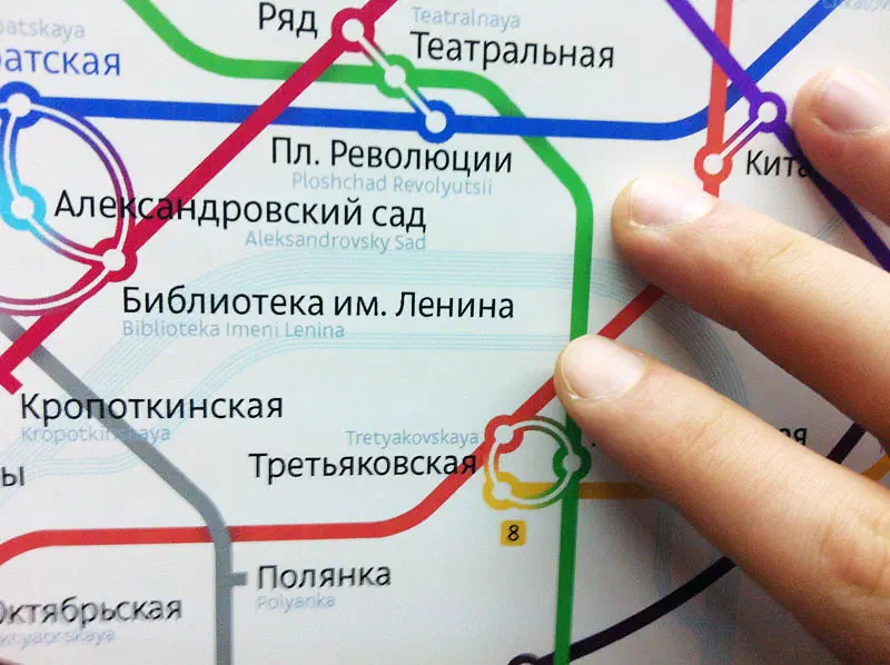 На картинке изображен человек, указывающий на карту системы метро с пальцем на ней. Карта заполнена красочными линиями и текстом, указывающим на различные остановки и маршруты системы метро.