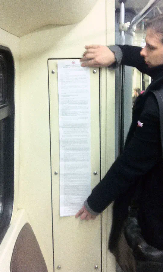 Изображение показывает, что человек стоит на поезде метро, держа кусок бумаги в руке. Он носит мешок и окно на левой стороне изображения.