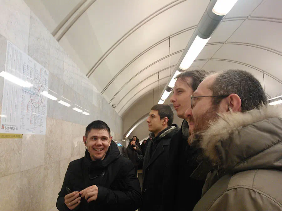 На снимке изображена группа людей, стоящих на станции метро, освещенная огнями, прикрепленными к потолку. На левой стороне изображения есть бумага, прикрепленная к стене с некоторым текстом на ней.