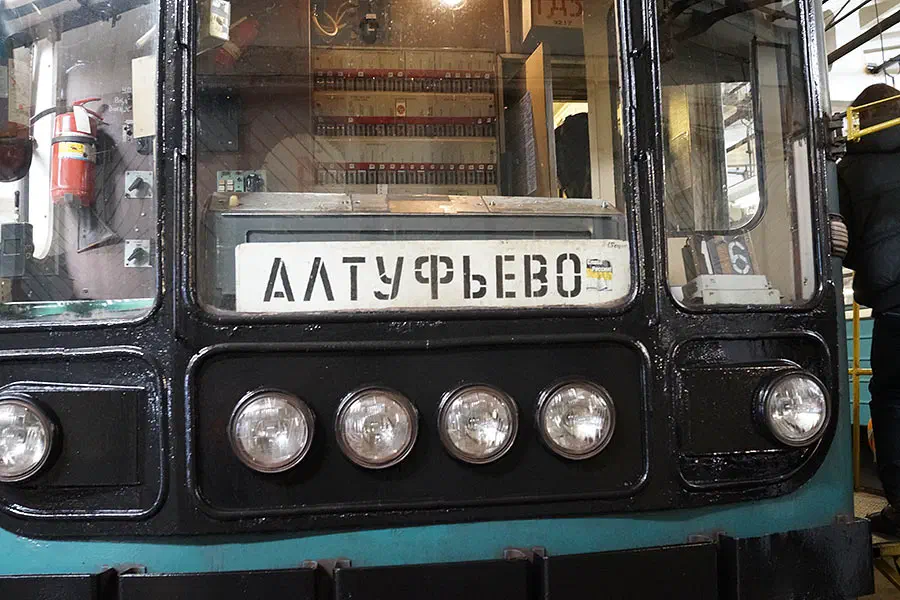 На снимке изображен человек, стоящий рядом с голубым и черным автобусом в здании. У автобуса есть номерная пластина с текстом, а на левой стороне изображения есть огнетушитель.