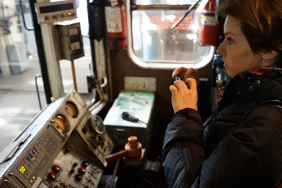Изображение показывает женщину, сидящую у управления поезда, держащую телефон в руке, окруженную огнетушителем, стеклянными окнами и другими предметами.