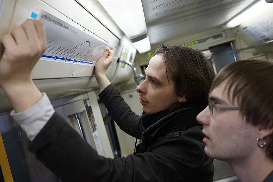 На снимке два человека смотрят на карту на поезде метро. Один из них держит бумагу в руке, а другой смотрит на карту. На потолке есть свет и плакаты.