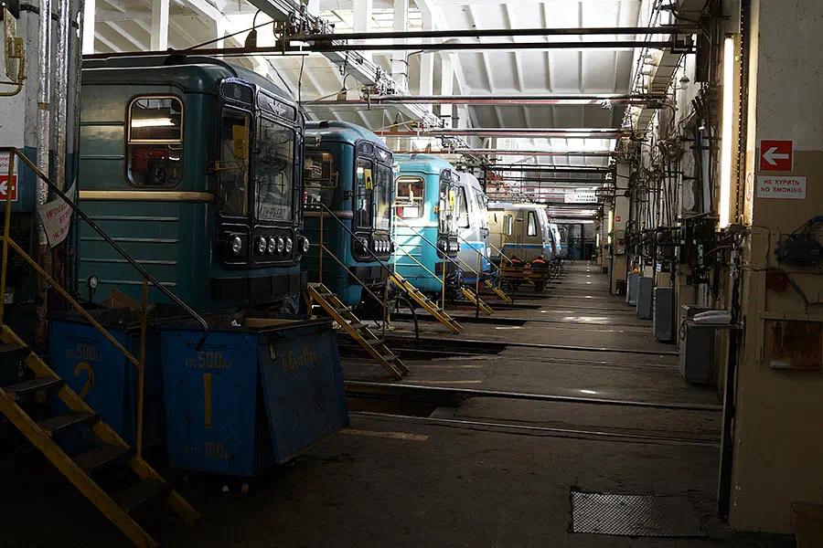Картина показывает большую комнату, заполненную множеством поездов, припаркованных внутри здания. Здесь есть ступеньки с перилами, ведущими к входу в здание, и щит с текстом на стене справа.