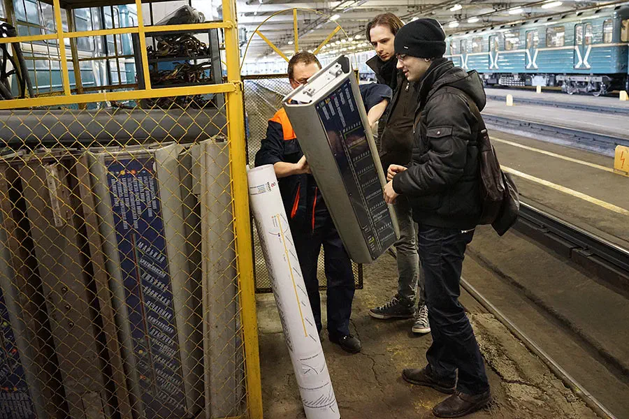 Изображается, что три человека стоят рядом друг с другом на станции метро, а поезд на железной дороге на заднем плане.