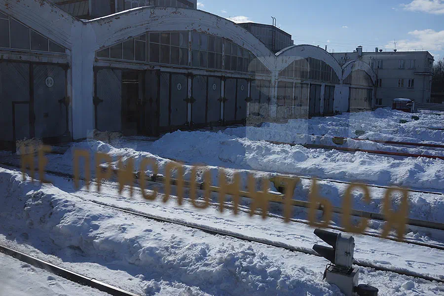 Изображение показывает железнодорожную станцию со снегом на земле и перед ней железнодорожный путь. Станция окружена зданиями с окнами и дверями, и на заднем плане деревья. Небо заполнено.