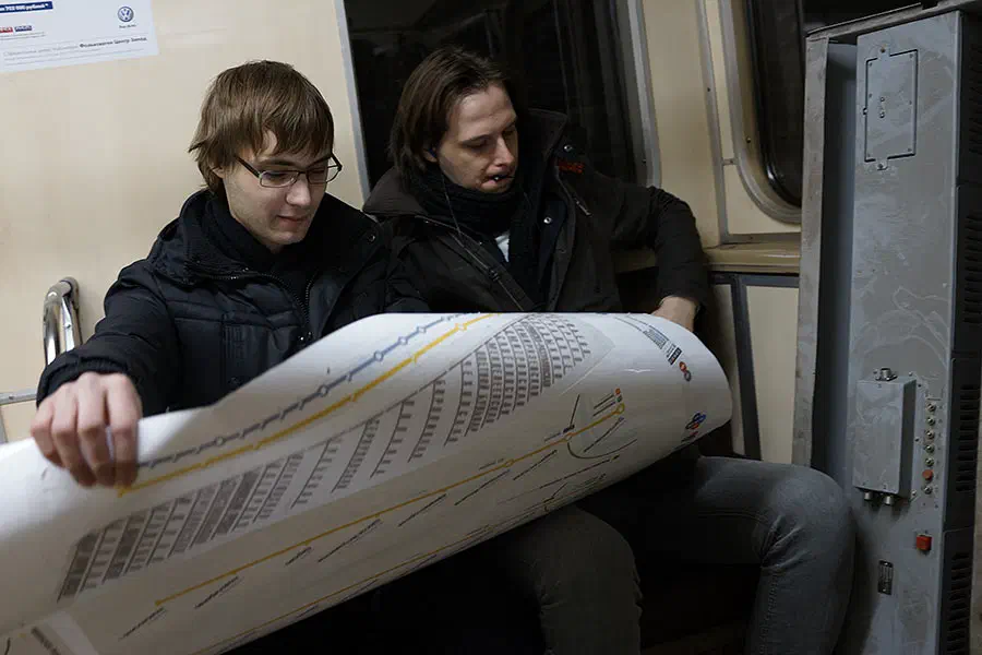 Изображение показывает, что два человека сидят в поезде метро, оба в черных пиджаках и целенаправленно смотрят на чертеж. Справа от изображения есть металлический объект, и слева, там.