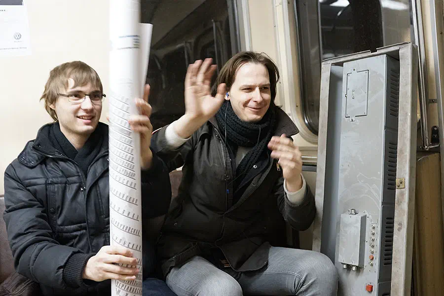 На снимке два человека сидят в поезде метро, оба в черных пиджаках и улыбаясь. Один из них держит бумагу в руке, а другой держит объект.