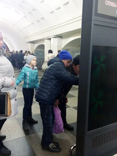 Изображение показывает группу людей, стоящих в станции метро рядом друг с другом, некоторые из которых держат мешки.
