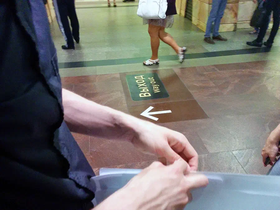 На рисунке изображена группа людей, стоящих на полу станции метро, и один человек держит объект в руке. На заднем плане есть стена со знаком "Bix"