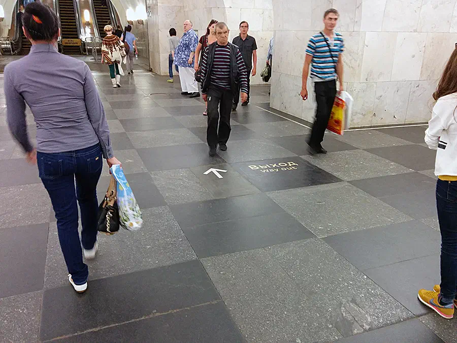На снимке изображена группа людей, проходящих через станцию метро, некоторые из которых носят мешки. На заднем плане есть эскалаторы, свет и стена, а некоторые из них носят джинсы.