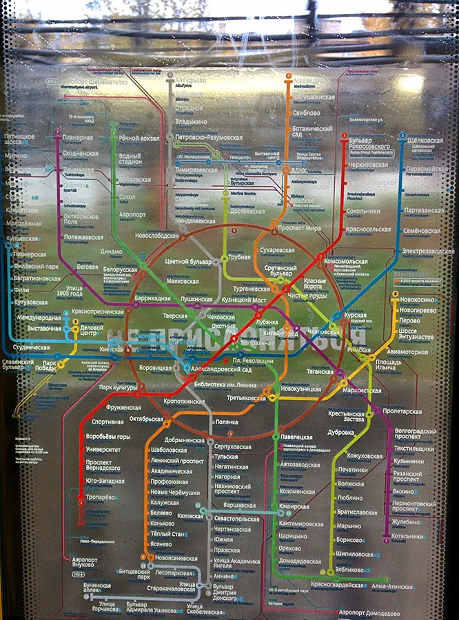 Карта показывает карту московской системы метро на стеклянном окне с деревьями на заднем плане. Карта представляет собой подробное представление системы метро с четко видимыми линиями и станциями.