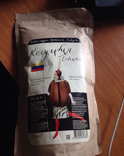 Картина показывает пакет кофе, сидящий на деревянном столе, с бумажной обложкой с изображением дамы и текстом на ней, а также проволокой с правой стороны.