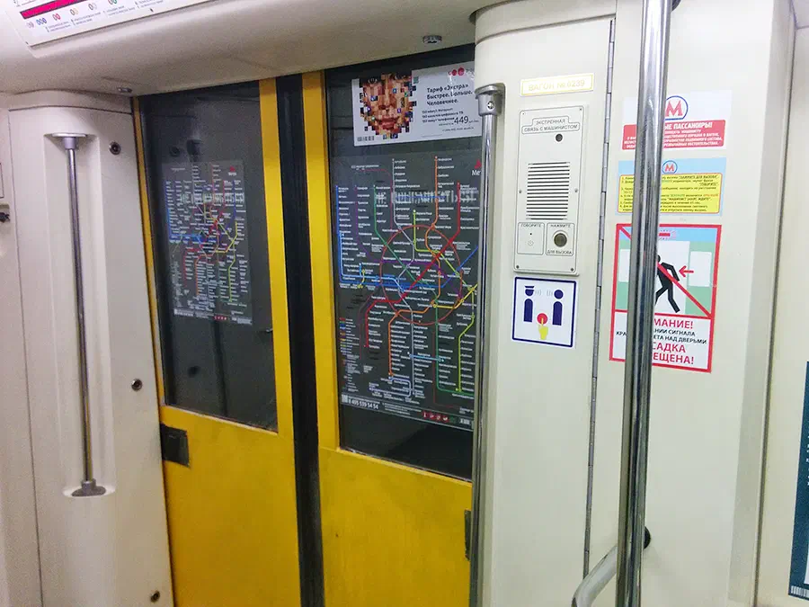 Изображение показывает поезд метро с желтой дверью и картой на стене. Дверь украшена плакатами, и есть ручки по обе стороны двери.
