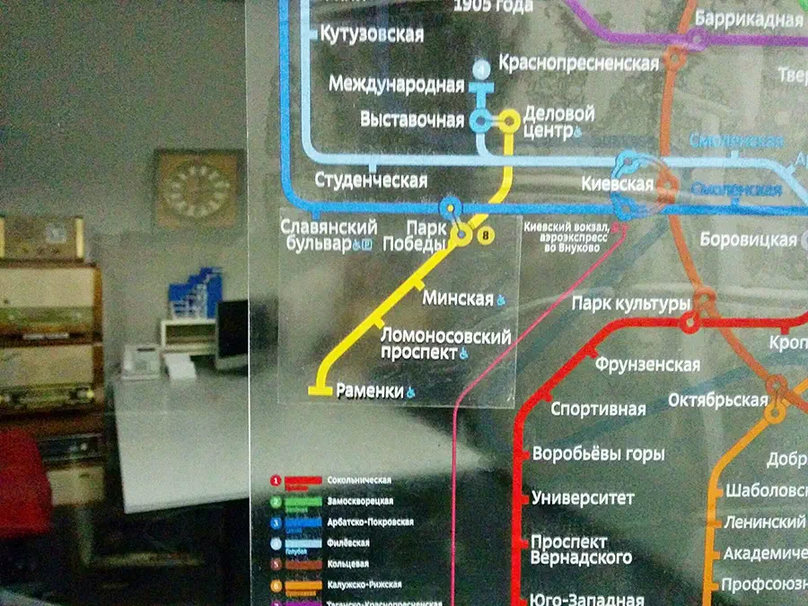 Изображение показывает карту системы метро на экране компьютера в комнате со стеной на заднем плане. На левой стороне изображения есть стол с различными предметами, включая часы.