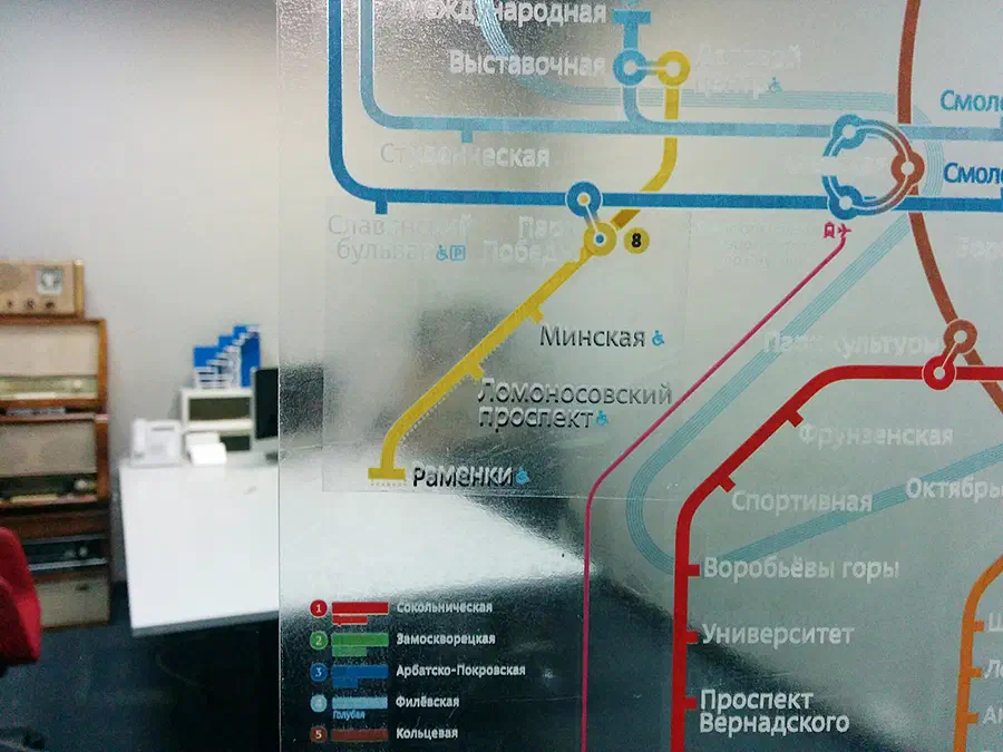 Карта московской системы метро на стеклянной стене в офисе. На левой стороне изображения есть стул и стол с различными предметами, включая монитор и другие объекты.