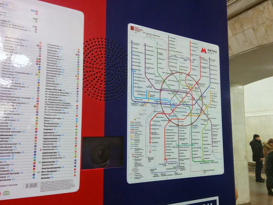 Изображение показывает плакат на стене станции метро с картой московской системы метро, несколько человек стоят на полу справа от плаката, и стена на стене.