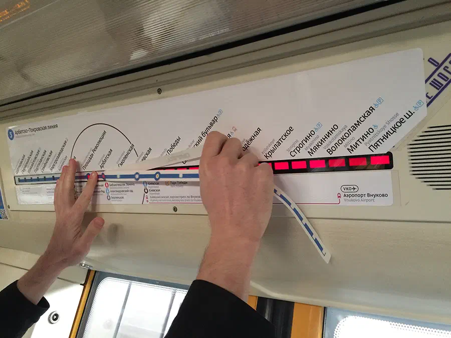 На рисунке изображен человек, держащий кусок бумаги в руке, указывая на карту в поезде метро. Руки этого человека видны, а карта прикреплена к потолку поезда с окнами.