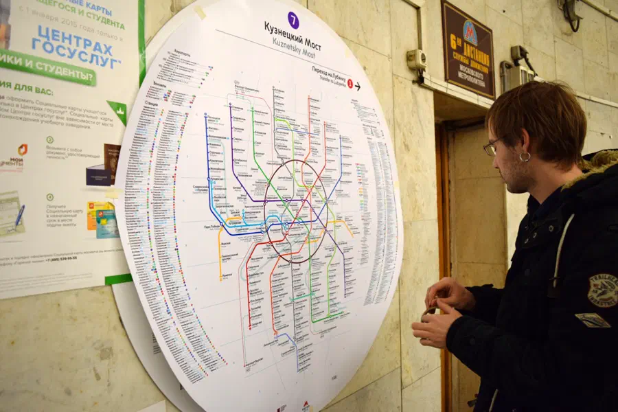 На снимке изображен человек, стоящий перед стеной с картой московской системы метро, он внимательно смотрит на карту, которая прикреплена к стене с плакатами и другими предметами.