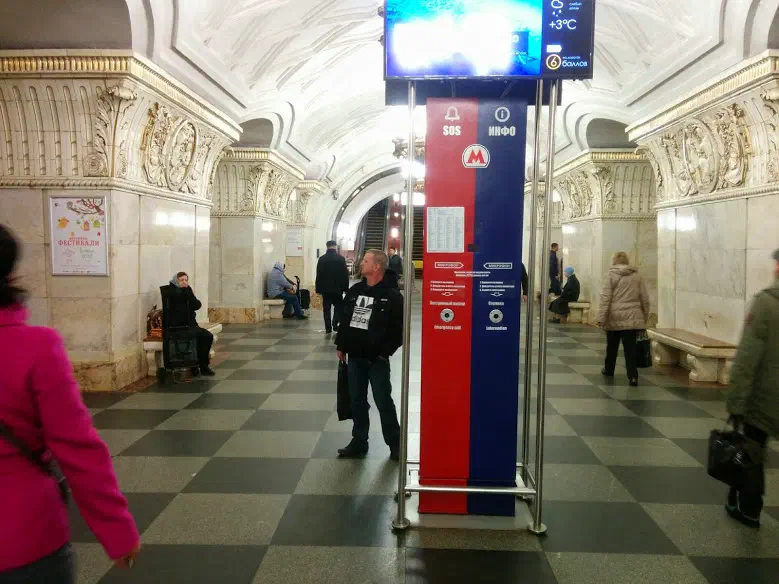 На картинке изображена группа людей, проходящих через станцию метро с большим экраном на стене: люди сидят на скамейках, некоторые стоят на полу, и плакат на левой стороне изображения.