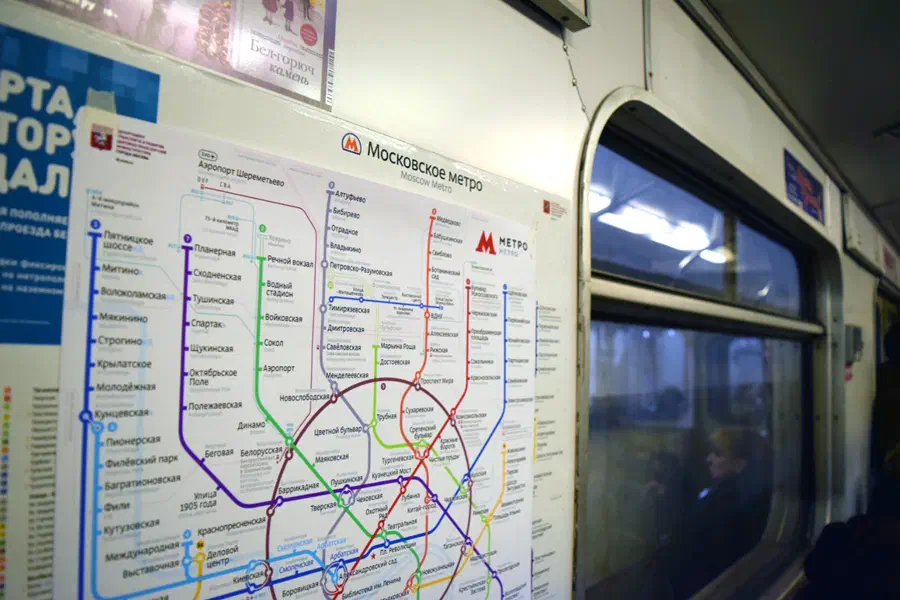 Изображение показывает поезд метро с картой московской системы метро на стене, плакаты с левой стороны изображения, стеклянное окно с правой стороны и несколько человек внутри поезда.